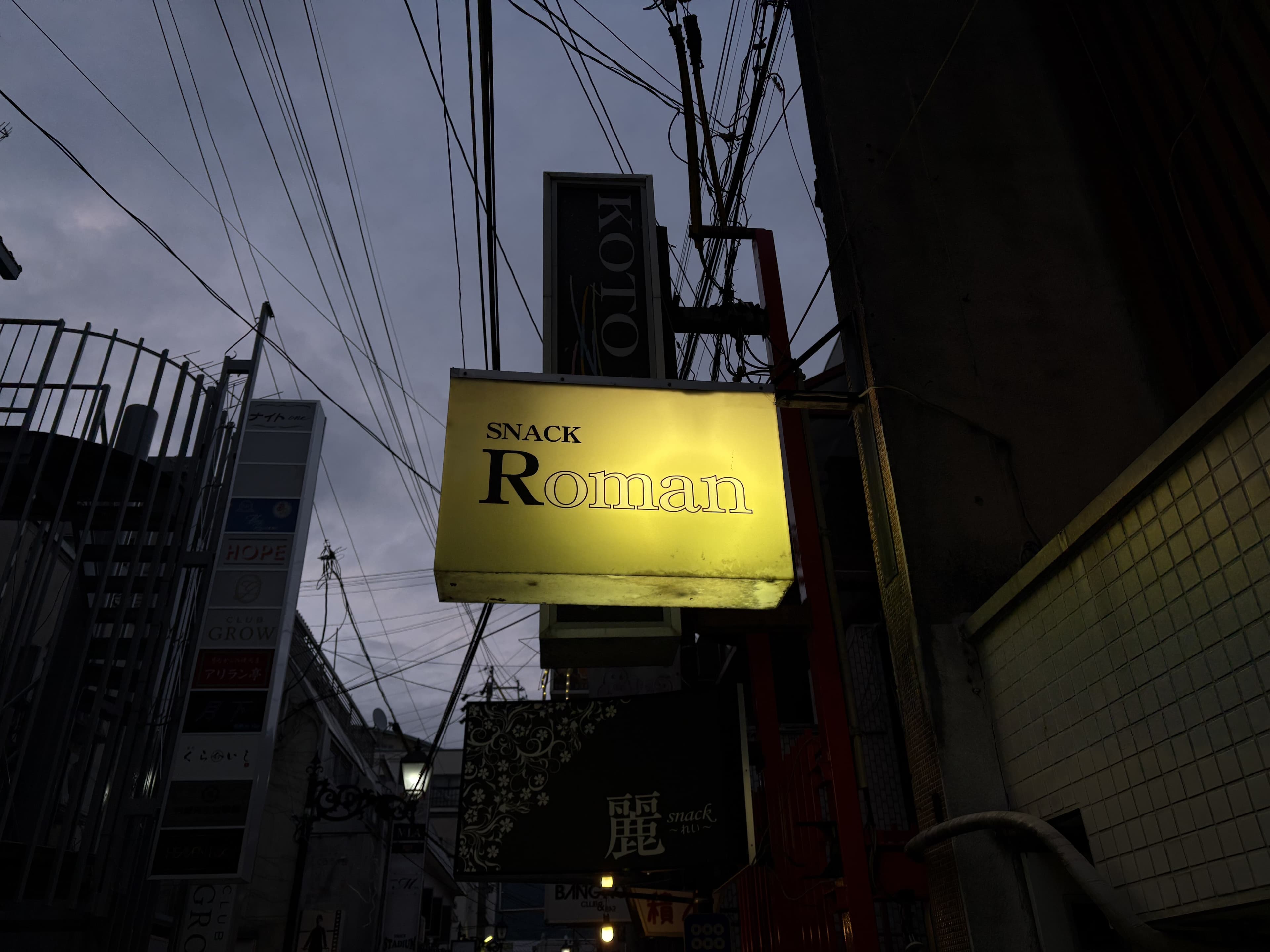 灯る黄色い「SNACK Roman」看板の路地風景